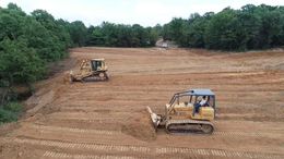 Two yellow bulldozers grading dirt in a cleared field near a tree line.