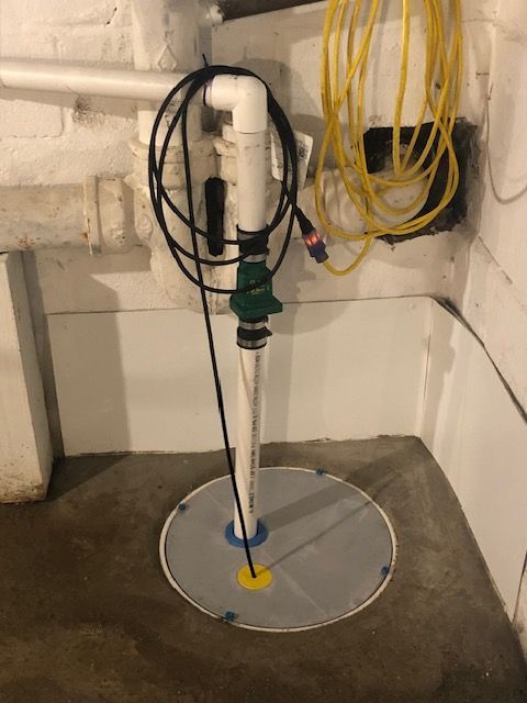 Basement sump pump with white pipe, coiled black and yellow hoses, and concrete floor drain cover