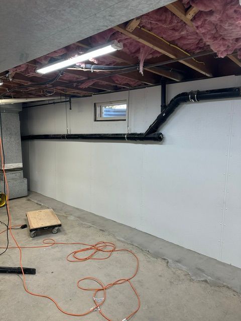 Basement under construction with white wall, exposed ceiling insulation, black ductwork, and extension cords on concrete floor