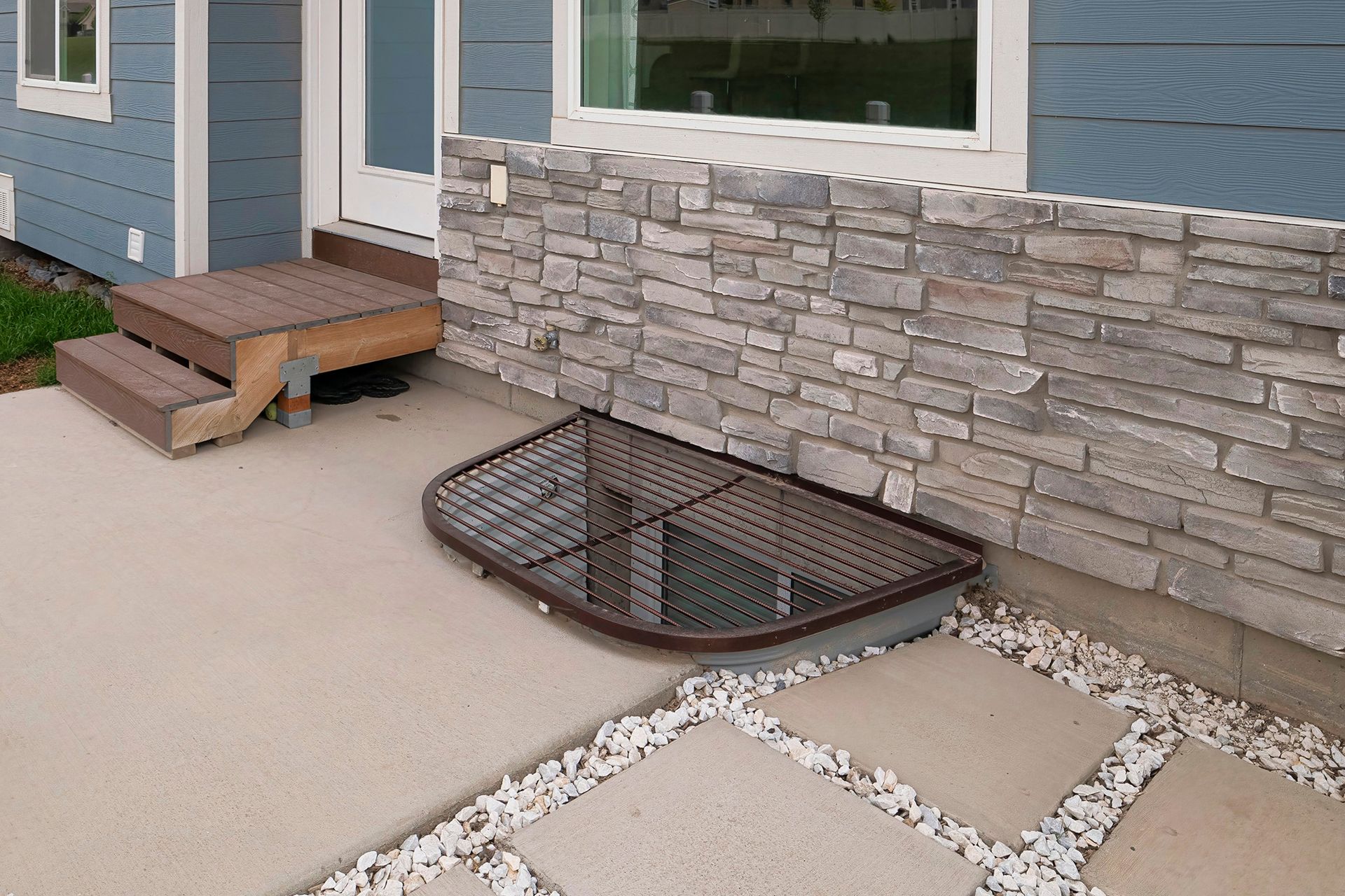 Basement window well beside stone-sided house with wood steps and a small concrete patio area.