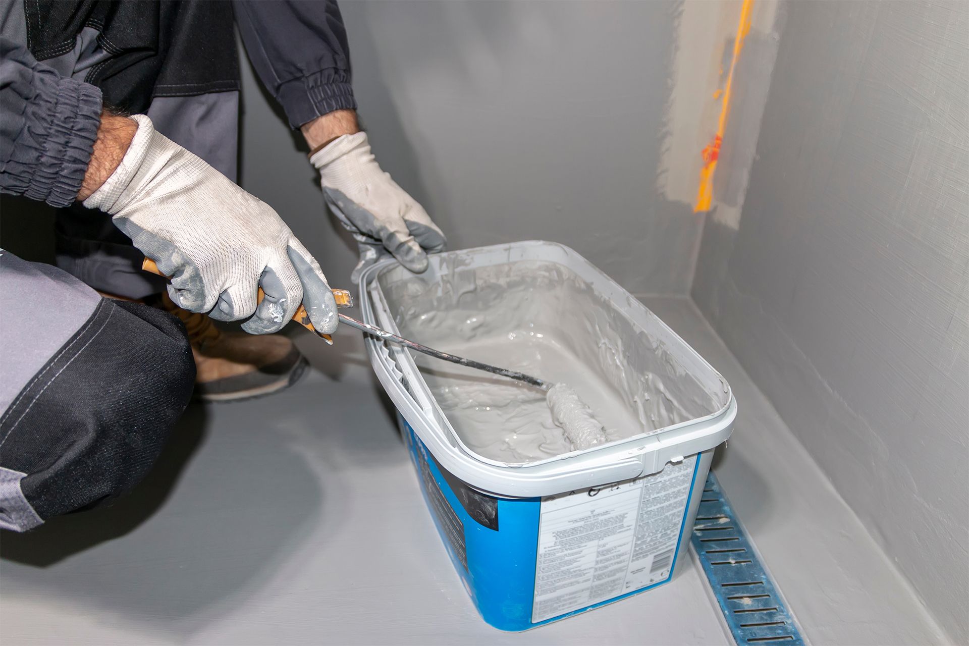Gloved hands mixing gray coating in a blue paint tray on a floor with a trowel
