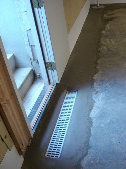 Interior floor with a white trench drain beside a doorway, showing a wet concrete surface.