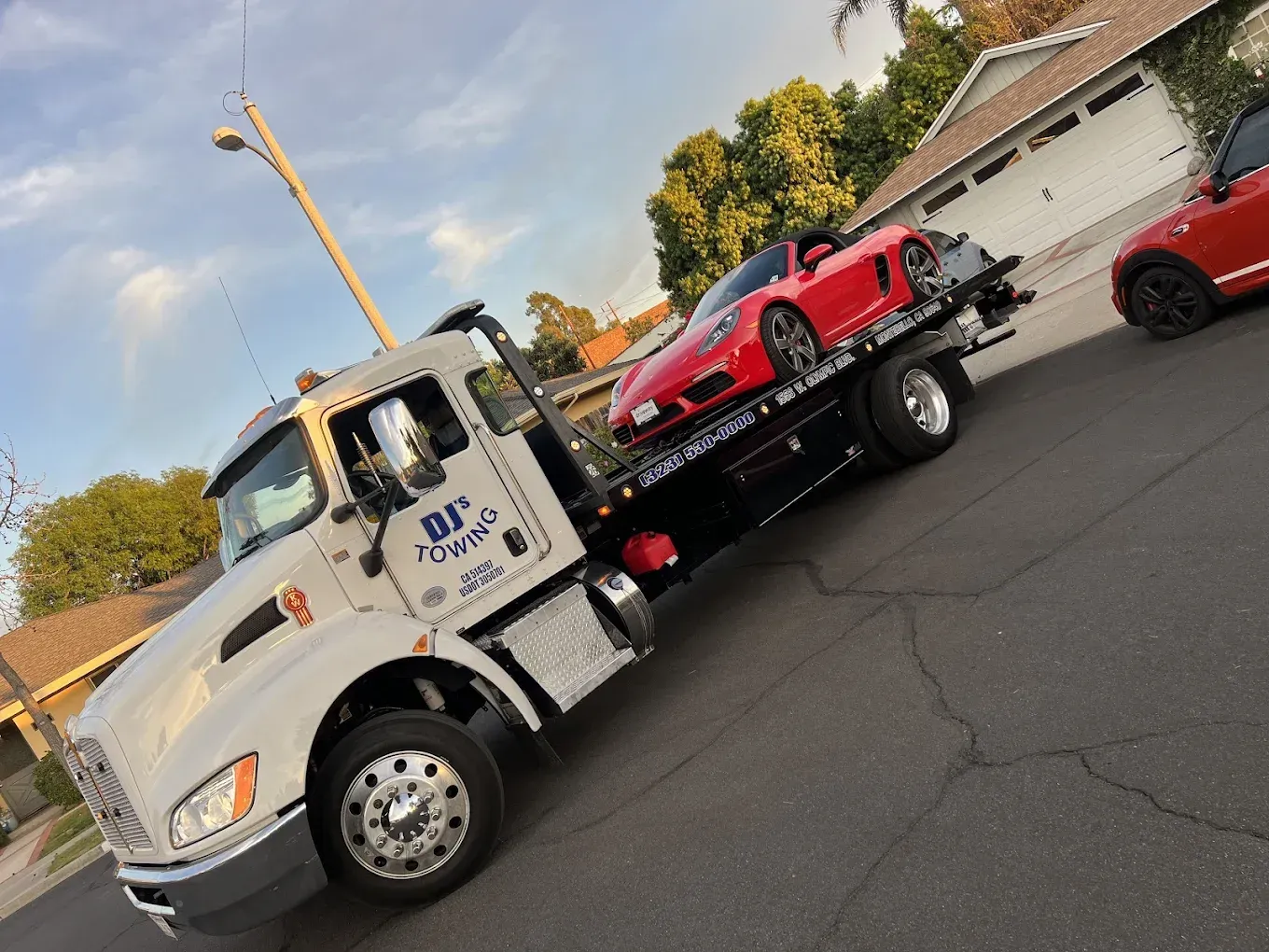 Tow truck towing a red car on a road. Truck is white with flashing lights.