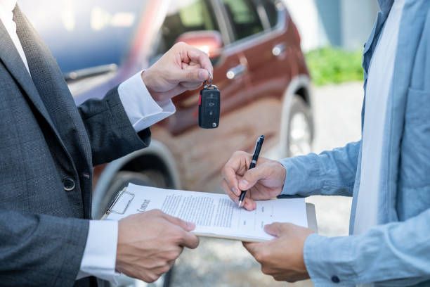 Person handing car keys to another person signing paperwork by a red car.