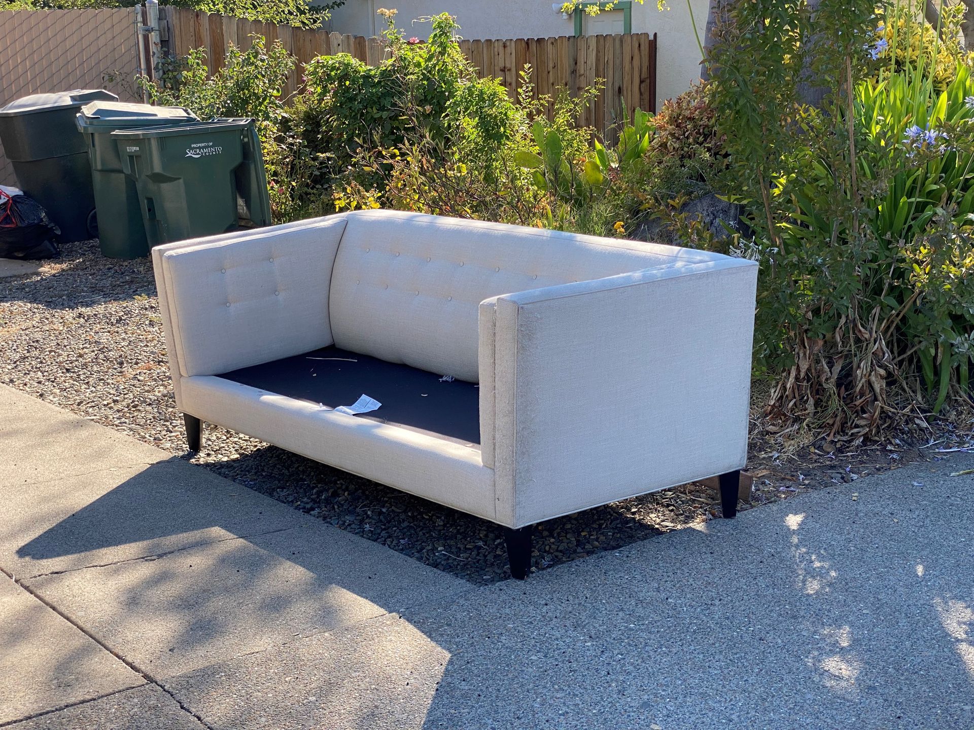 A white couch is sitting on a sidewalk next to trash cans.