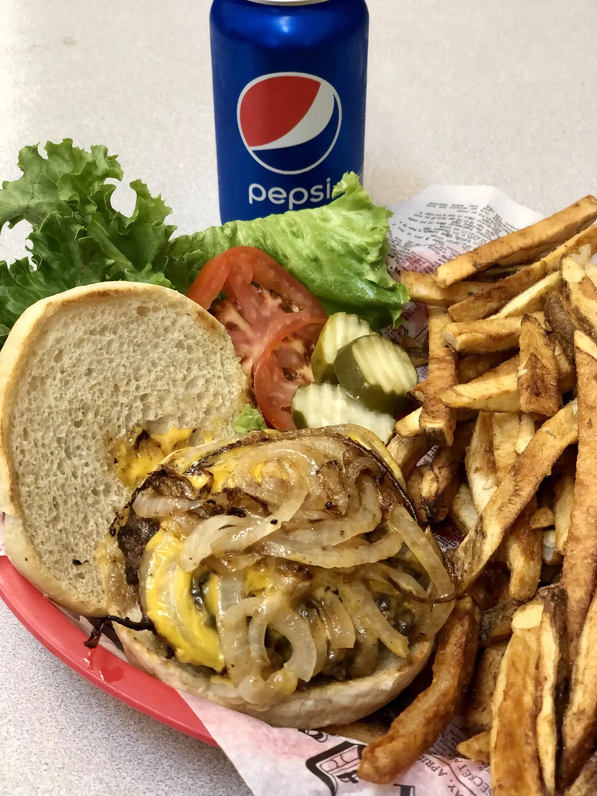 Burger with caramelized onions, fries, lettuce, tomato, pickles, and Pepsi can.