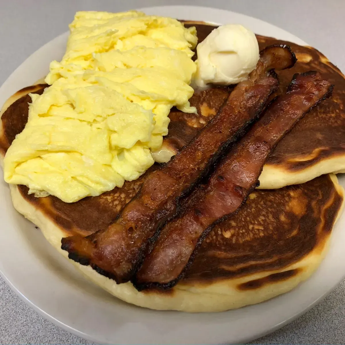 Plate of breakfast food: pancakes, scrambled eggs, bacon, and butter.