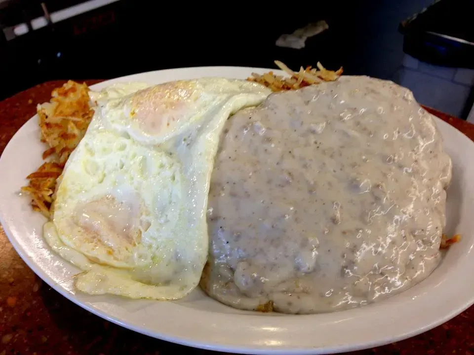 Plate of breakfast: sunny-side-up eggs, hash browns, and biscuit covered in sausage gravy.