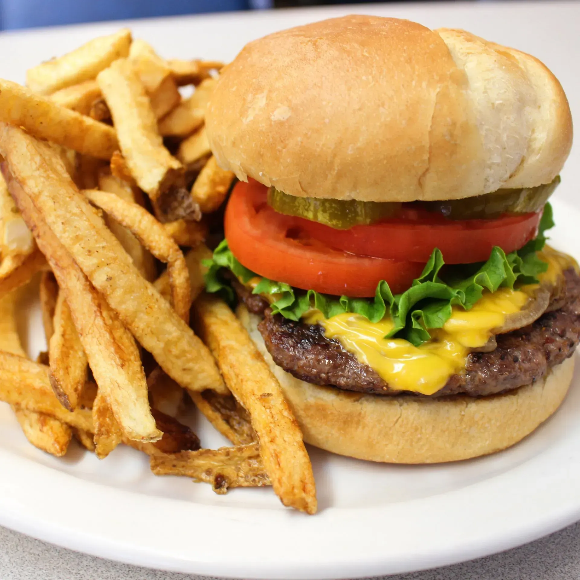 Cheeseburger with tomato, lettuce, and pickle on a bun, served with fries on a white plate.