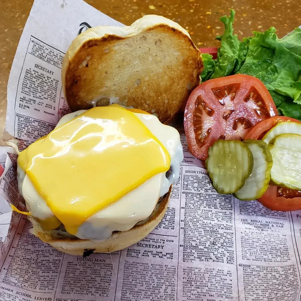Cheeseburger with melted cheese, tomato, pickles, lettuce, and toasted bun on printed paper.