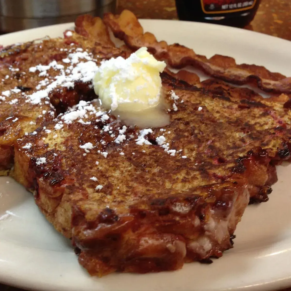 French toast with butter and powdered sugar, bacon on a white plate.