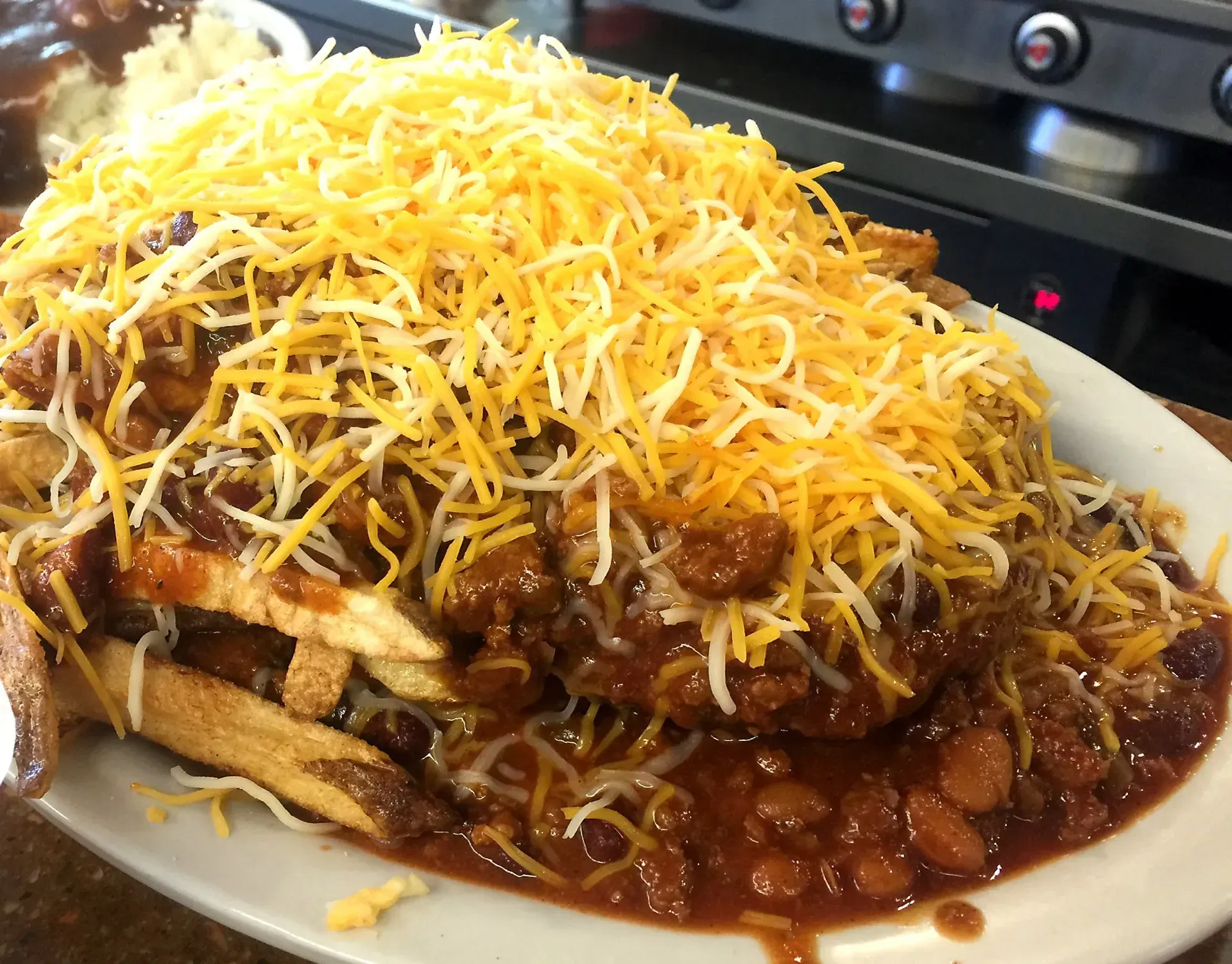 Fries, chili, and shredded cheddar cheese piled high on a white plate; kitchen setting.