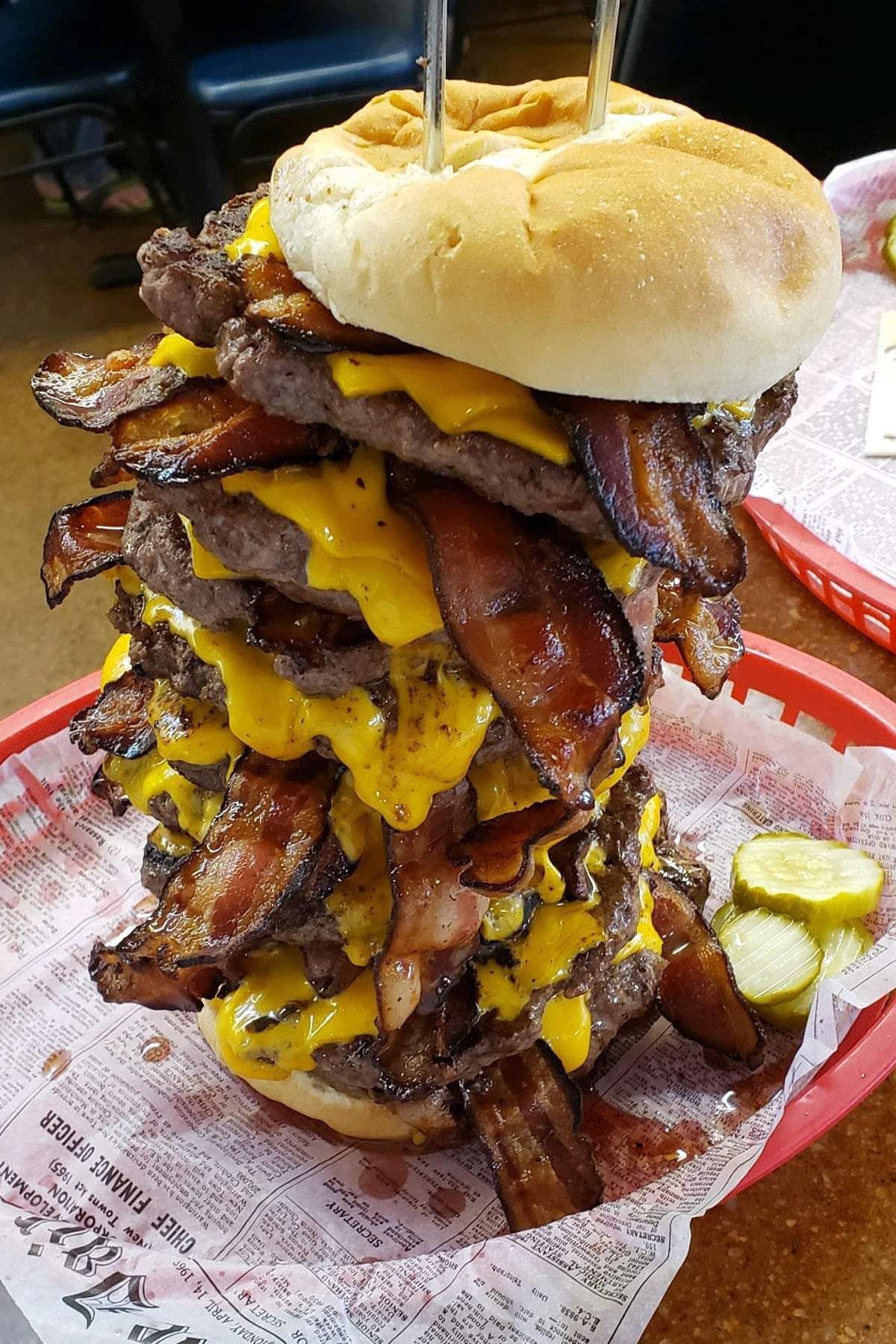 Towering burger with multiple patties, cheese, bacon, and pickles on a red tray.