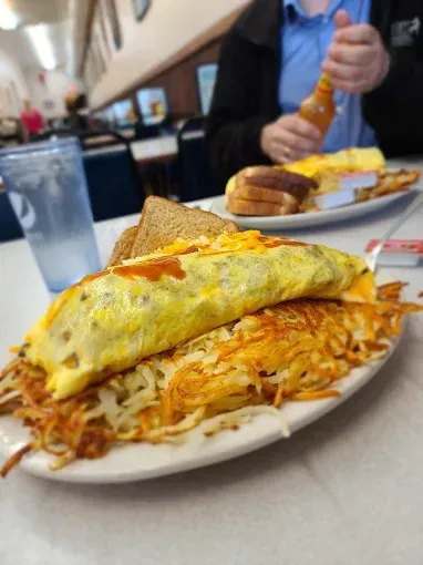 Omelet with hash browns and toast on a plate at a diner. A person adds hot sauce to a plate of eggs.