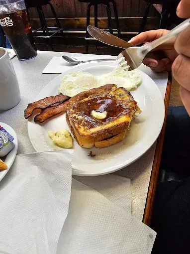 Breakfast plate: French toast with syrup and butter, eggs, bacon, and a hand holding a fork and knife.