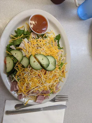 Salad with shredded cheese, cucumber slices, ham, and sauce in a small cup.