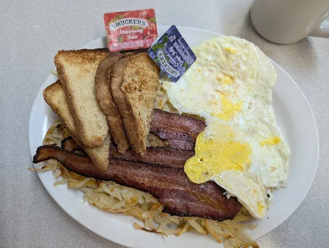 Breakfast plate: eggs, bacon, toast, hash browns, jelly packets.