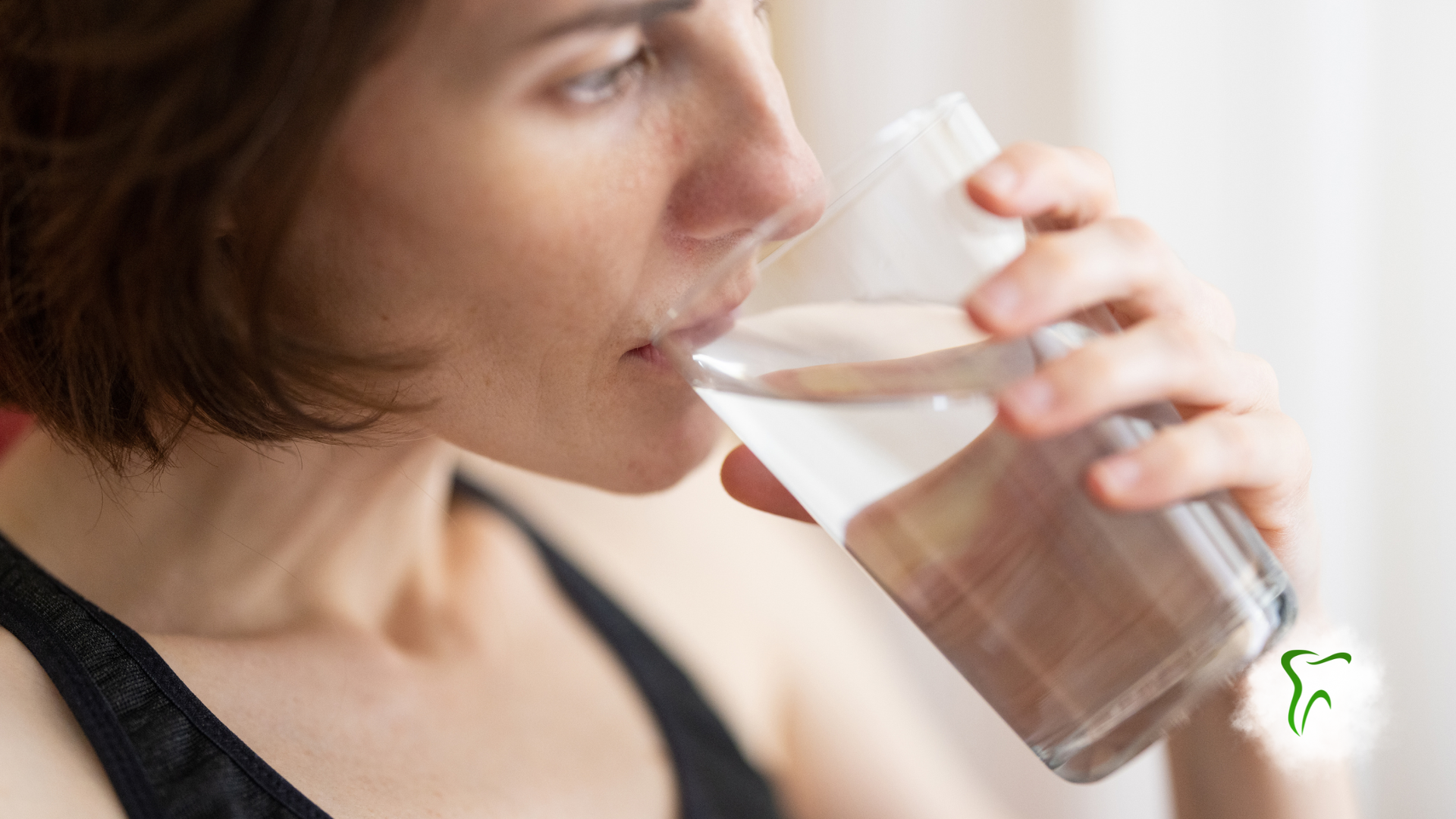 Woman drinking from a clear glass of water. Holding the glass with right hand, looking to the side.