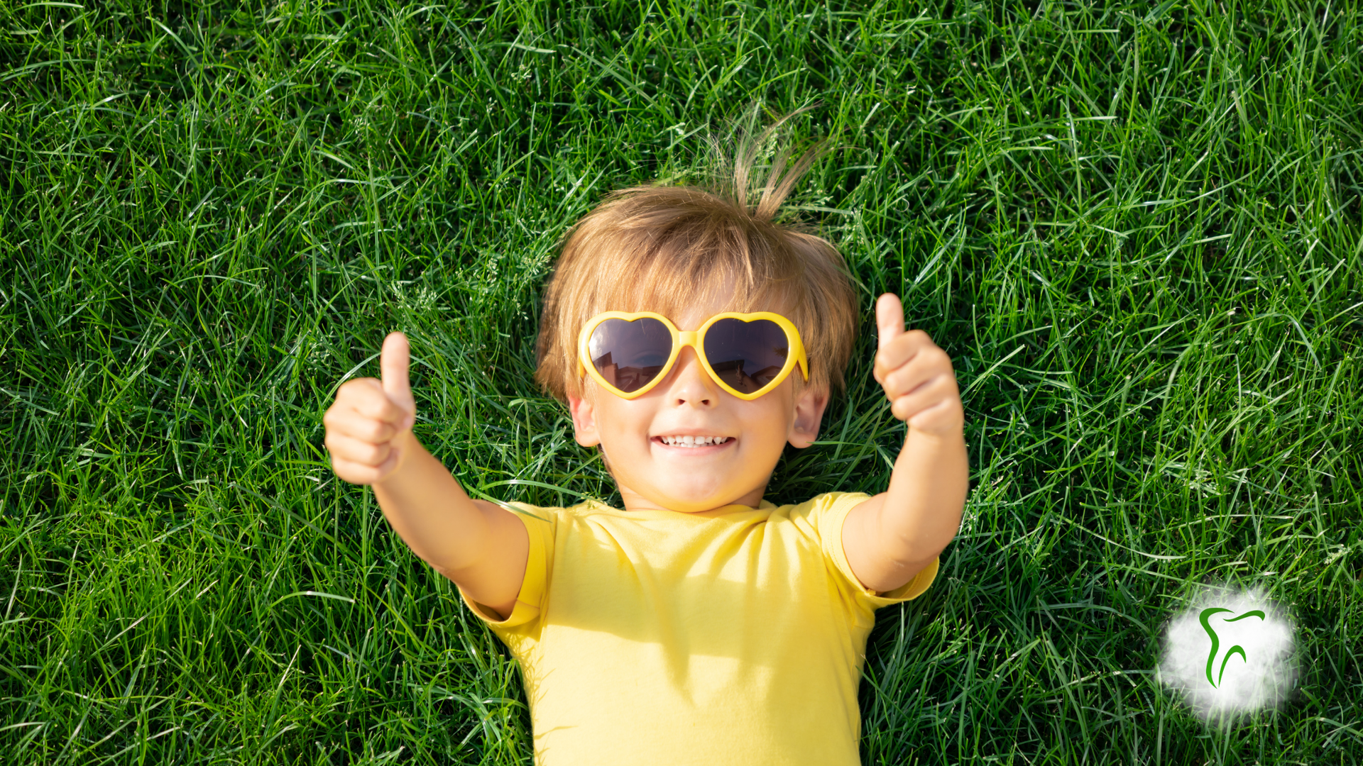 Child wearing heart-shaped sunglasses gives two thumbs up while laying in green grass.