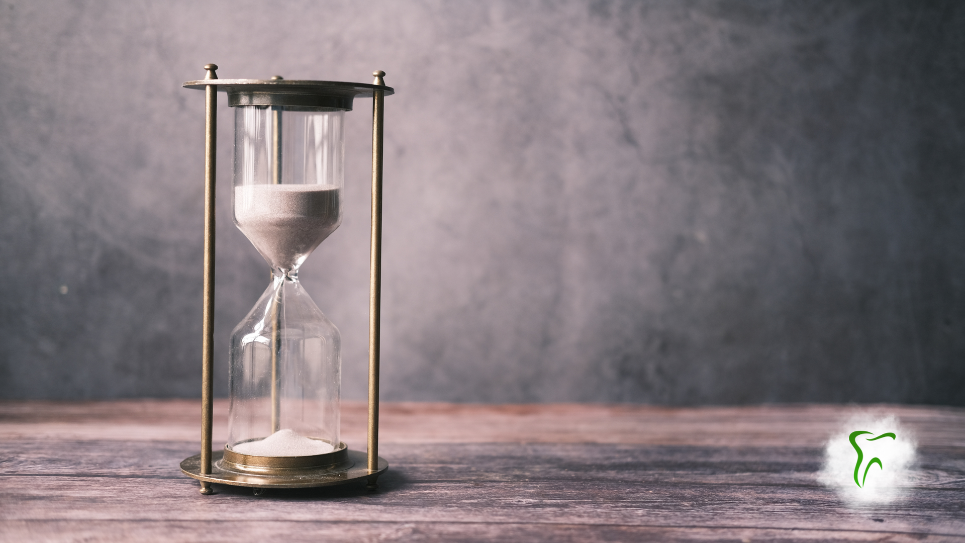 Hourglass on a wooden surface, sand flowing downwards. Dark gray background.