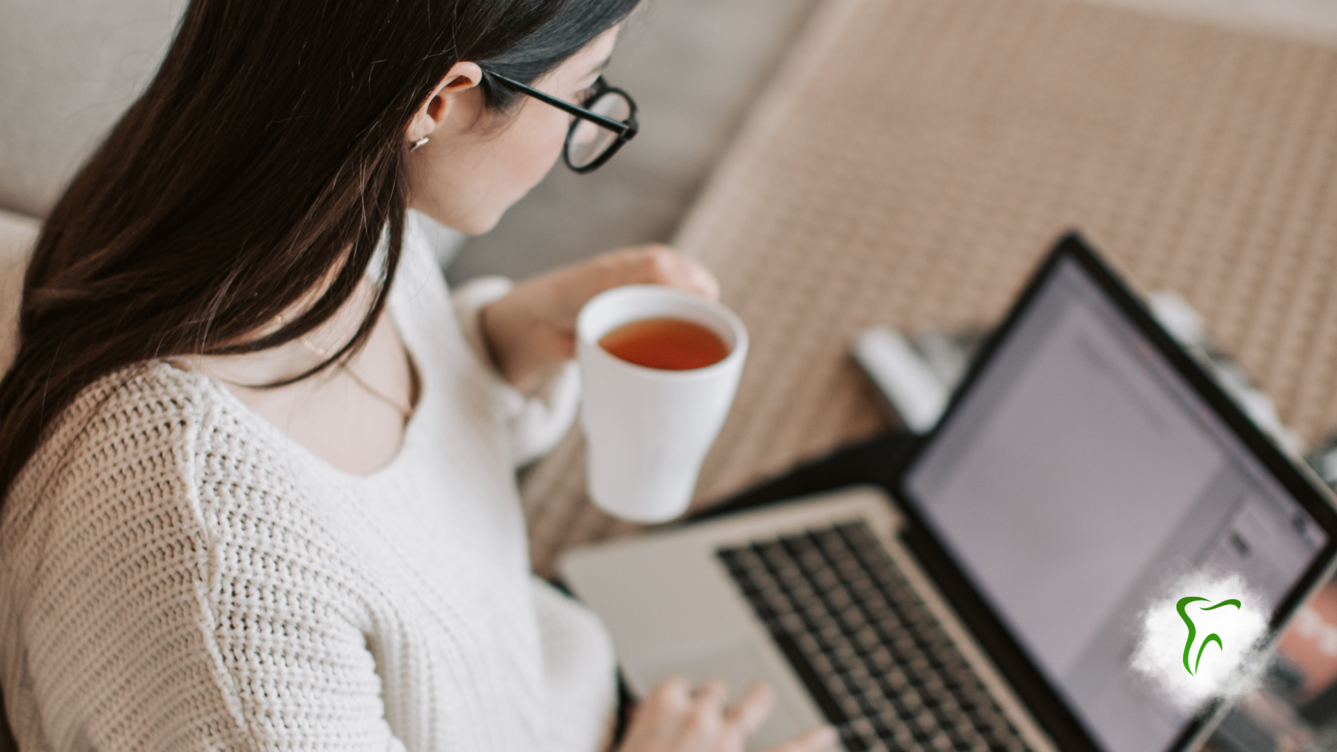 Woman in glasses and white sweater working on a laptop, holding a mug indoors.