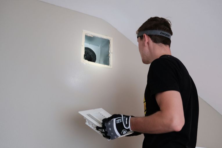 Person looking at square hole in wall; holding vent cover; wearing work gloves and headband.
