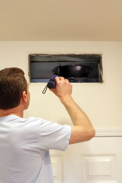 Man using a flashlight to inspect an open vent in a white wall.