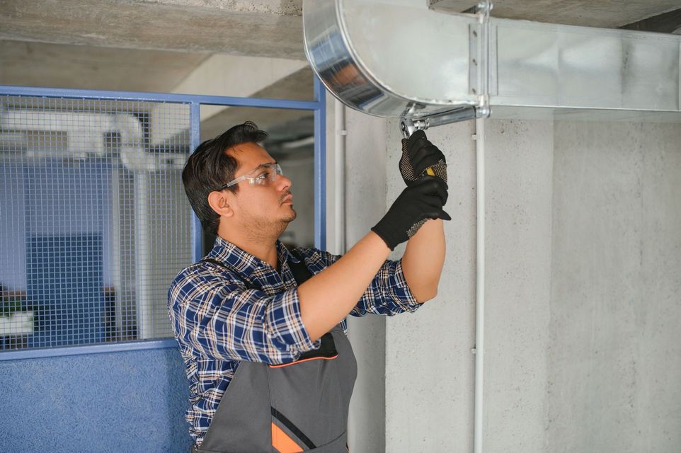 Man in work clothes and safety glasses works on HVAC ductwork in a workshop.