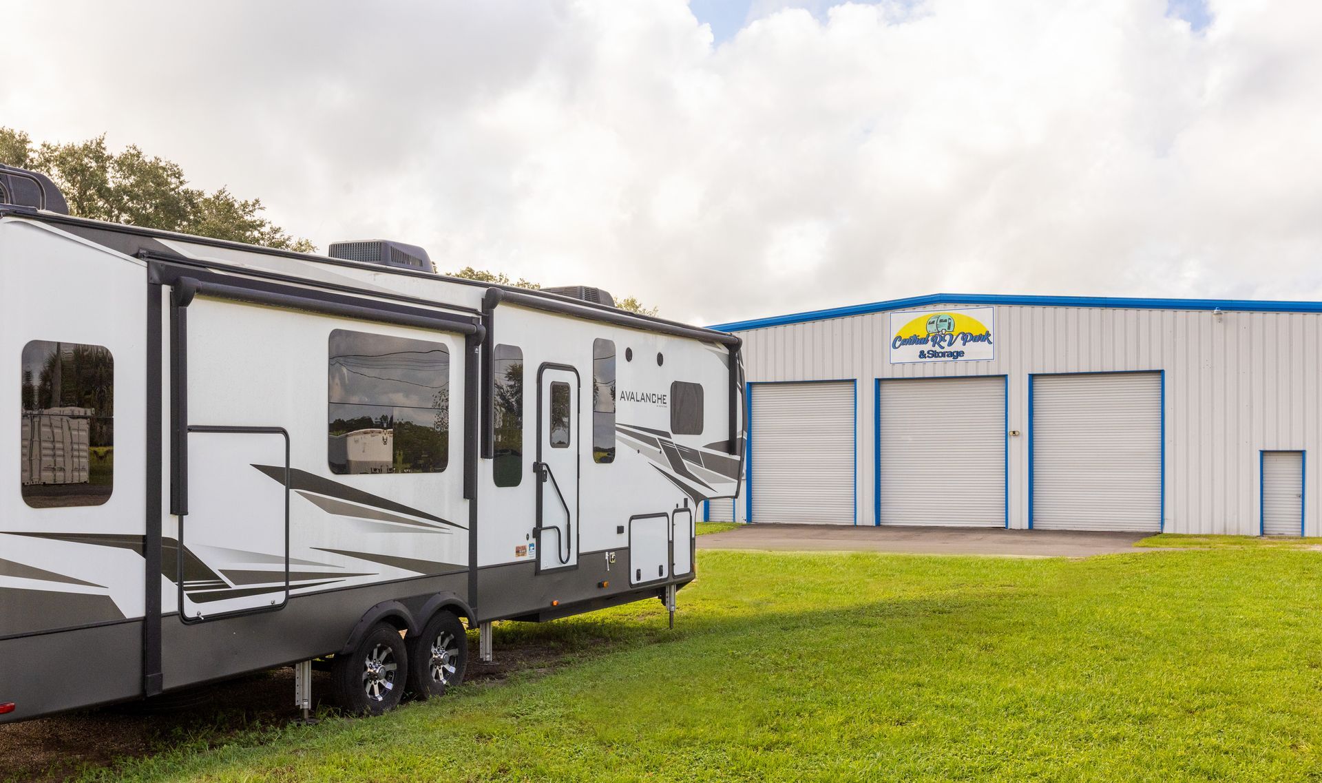 RV parked in front of a blue and white garage with business sign on a grassy field.