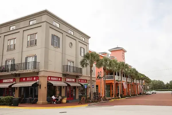 Buildings with shops on a brick street; palm trees, bright orange and grey facades, cloudy sky.