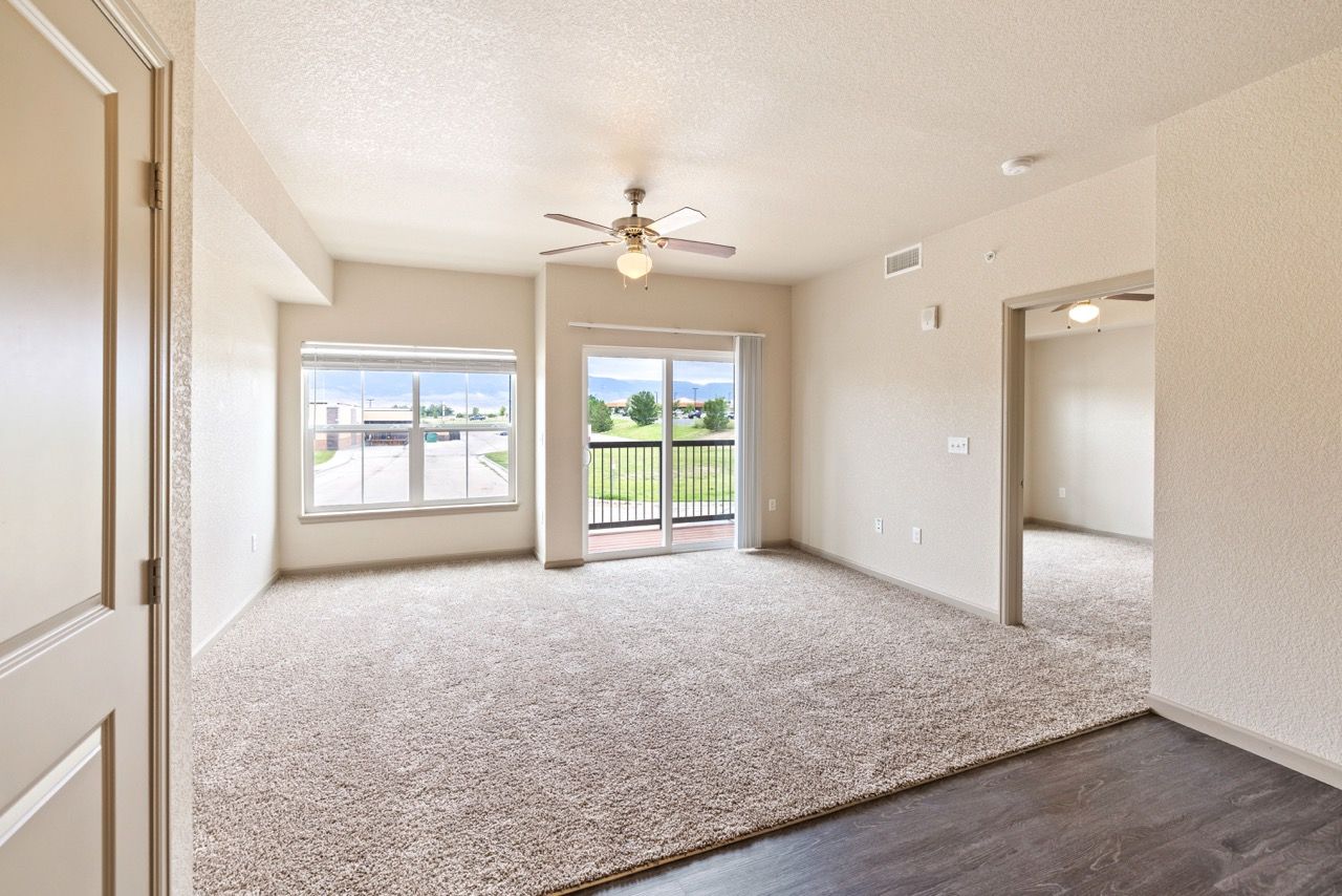 Living room with beige walls, carpet, ceiling fan, large window and sliding door to balcony.