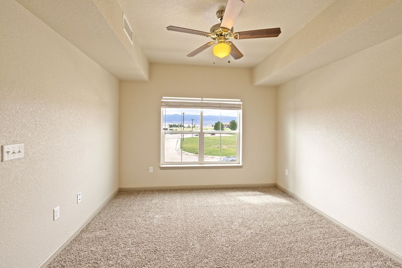 Empty beige carpeted room with a ceiling fan and a window showing outdoors.