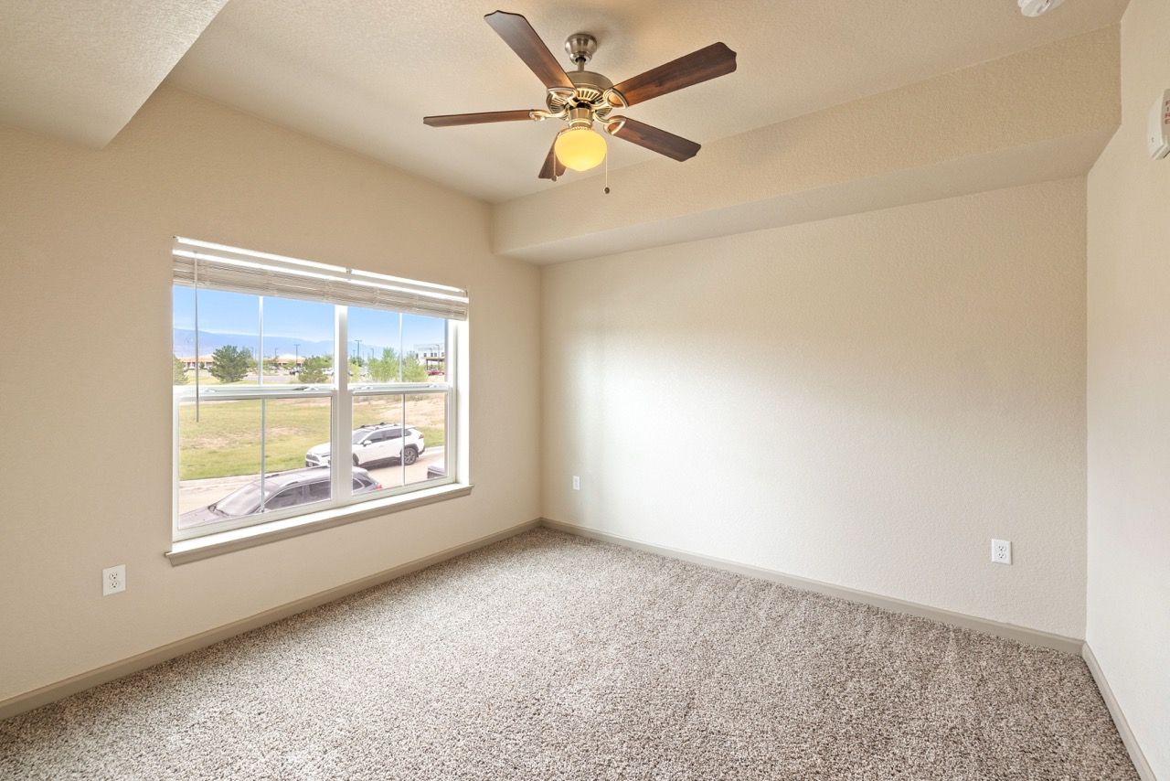 Vacant apartment bedroom with beige walls, carpet, a large window, and a ceiling fan.