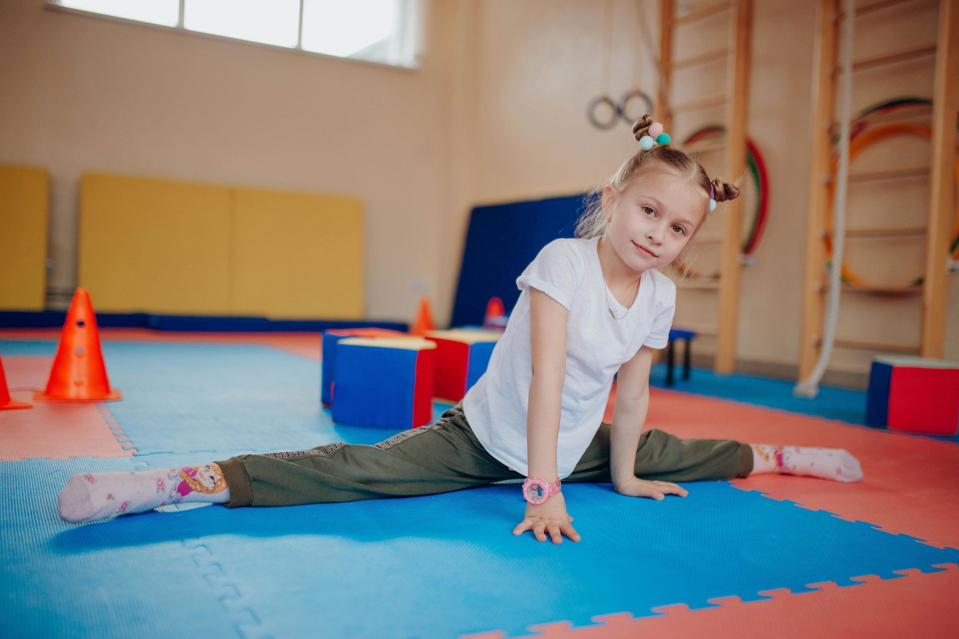 Child in a yellow shirt and purple pants sits on a yoga mat, stretching forward to hold her feet.