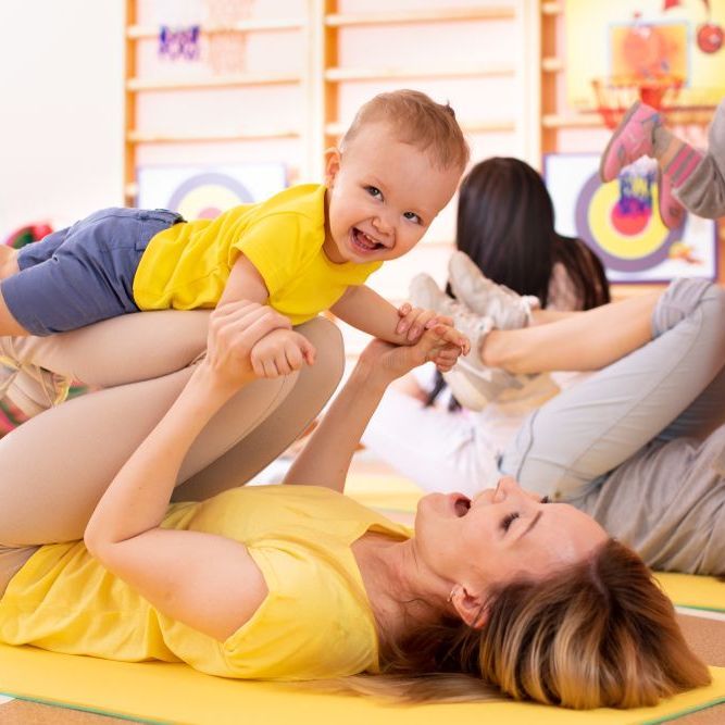 Woman in yellow shirt lifting a smiling baby in a bright playroom