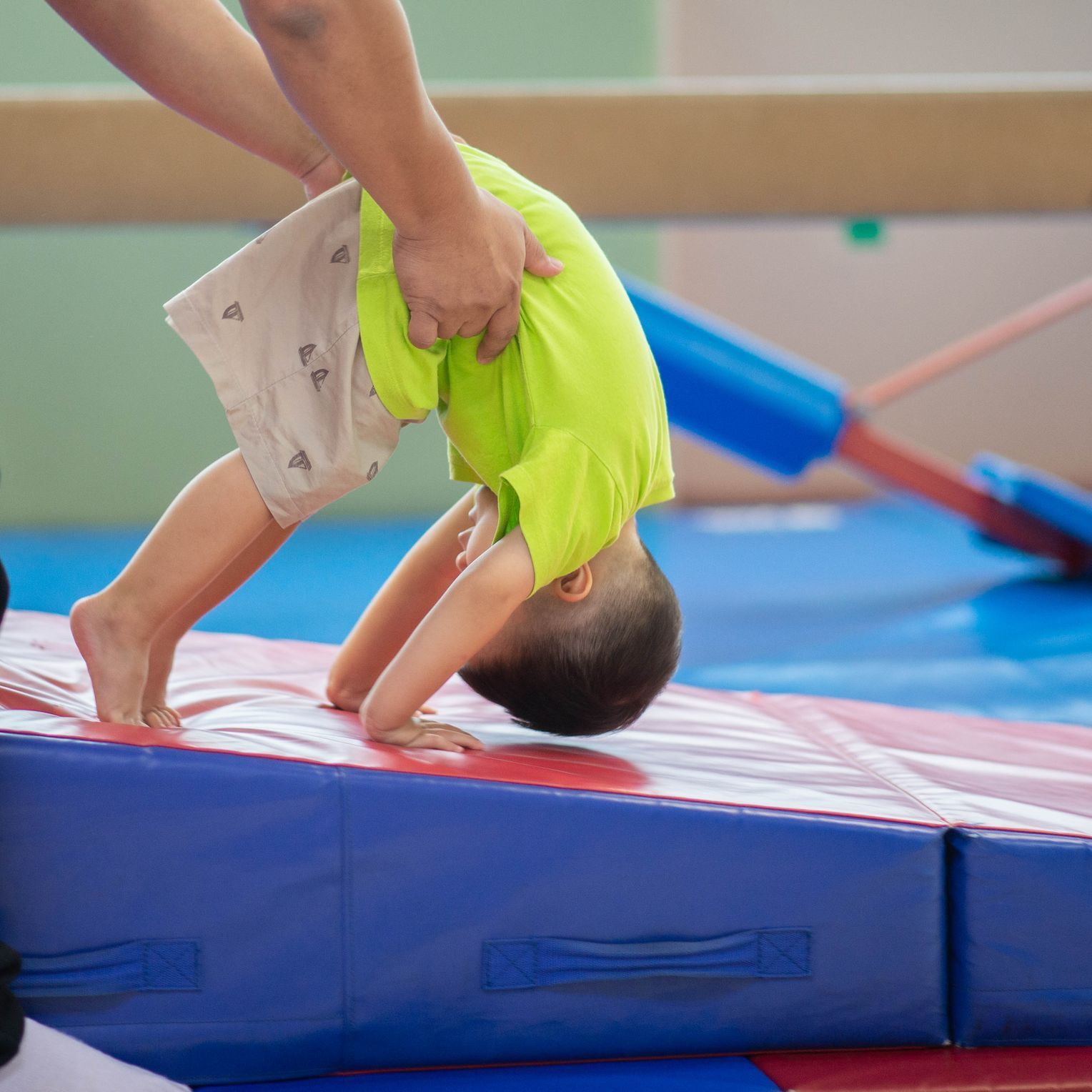Child doing a backbend on a red-and-blue gym mat with an adult assisting nearby