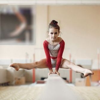 Gymnast in a red leotard balancing on a beam in a split pose, focused in a gym.