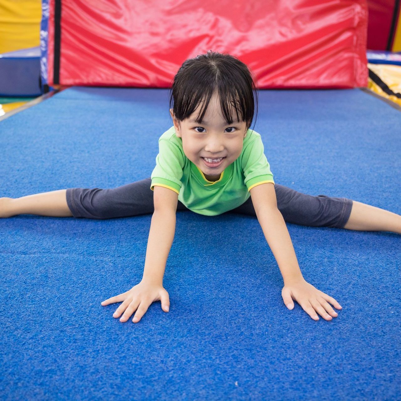 Child in a green shirt stretching on a blue mat in a gym, smiling toward the camera.