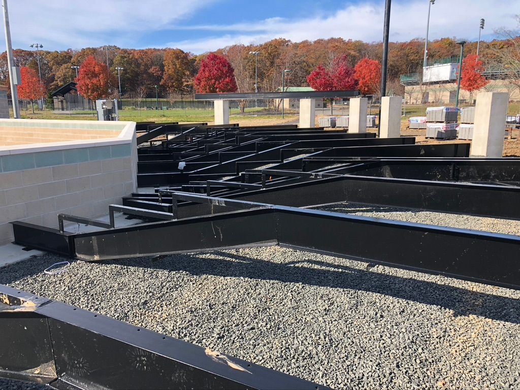 Construction site with black metal structures, gravel, and concrete features under a blue sky with fall foliage.