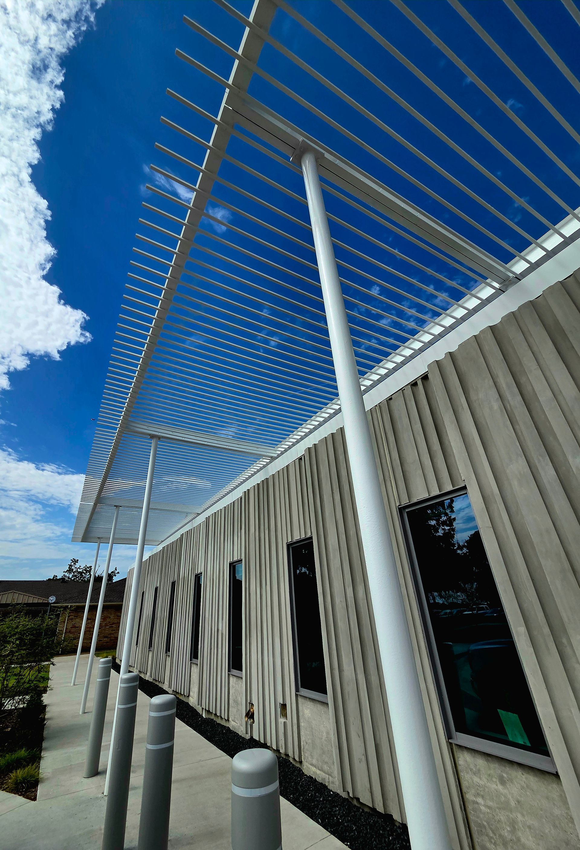 Exterior of a building with a white trellis, vertical siding, and windows; bright blue sky with clouds.