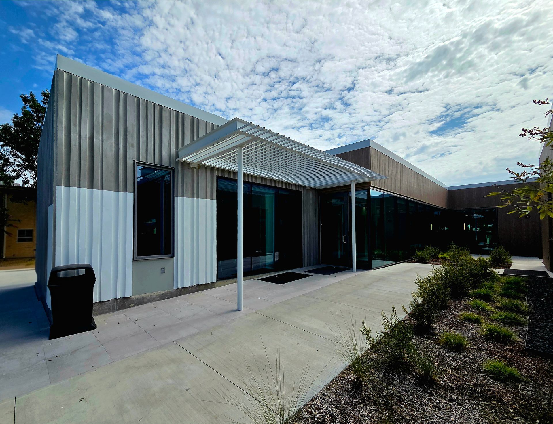 Modern building with corrugated metal siding, glass windows, and a shaded entrance under a partially cloudy sky.