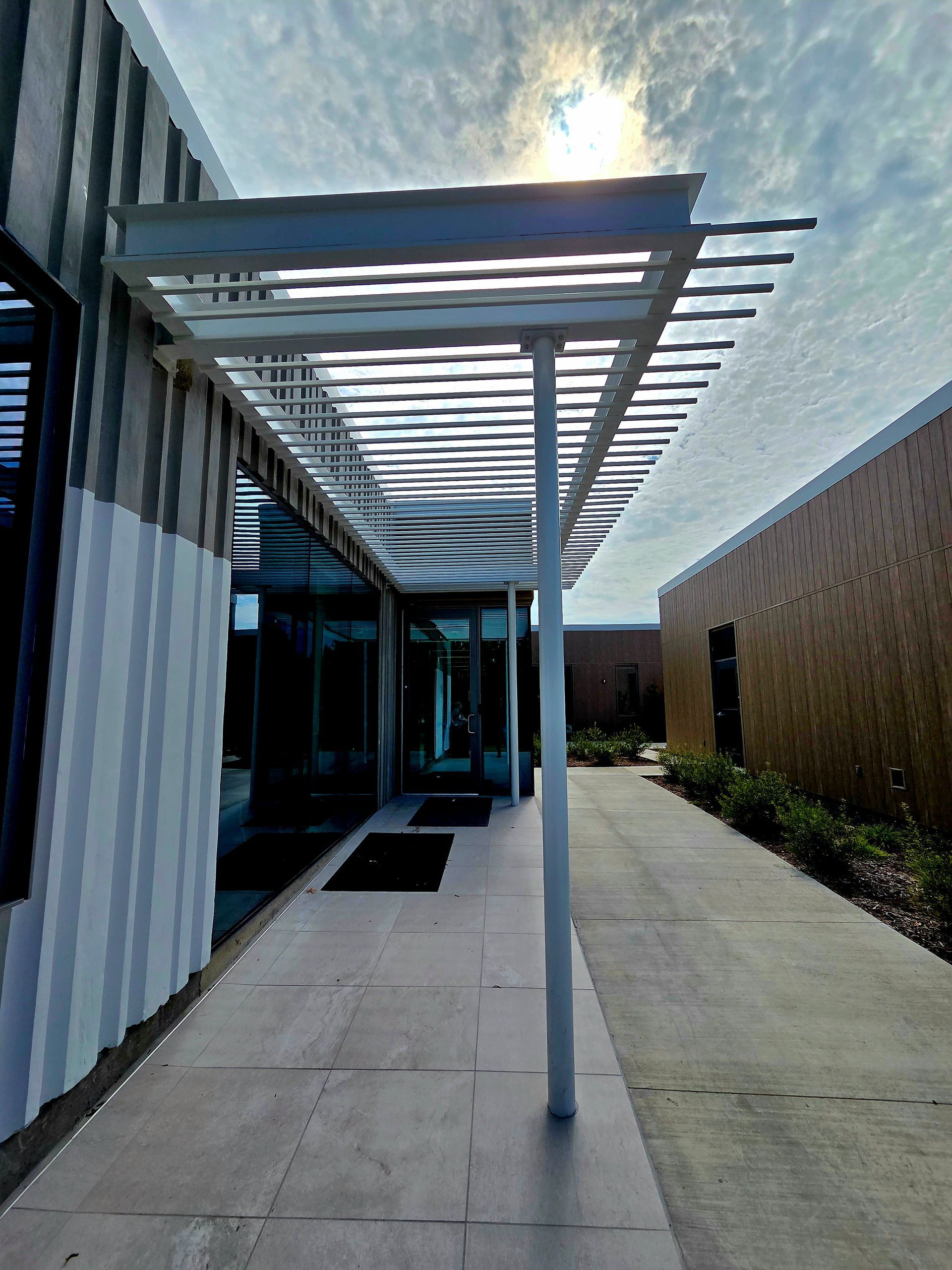 White pergola covers a walkway leading to a glass door; bright sunlight.