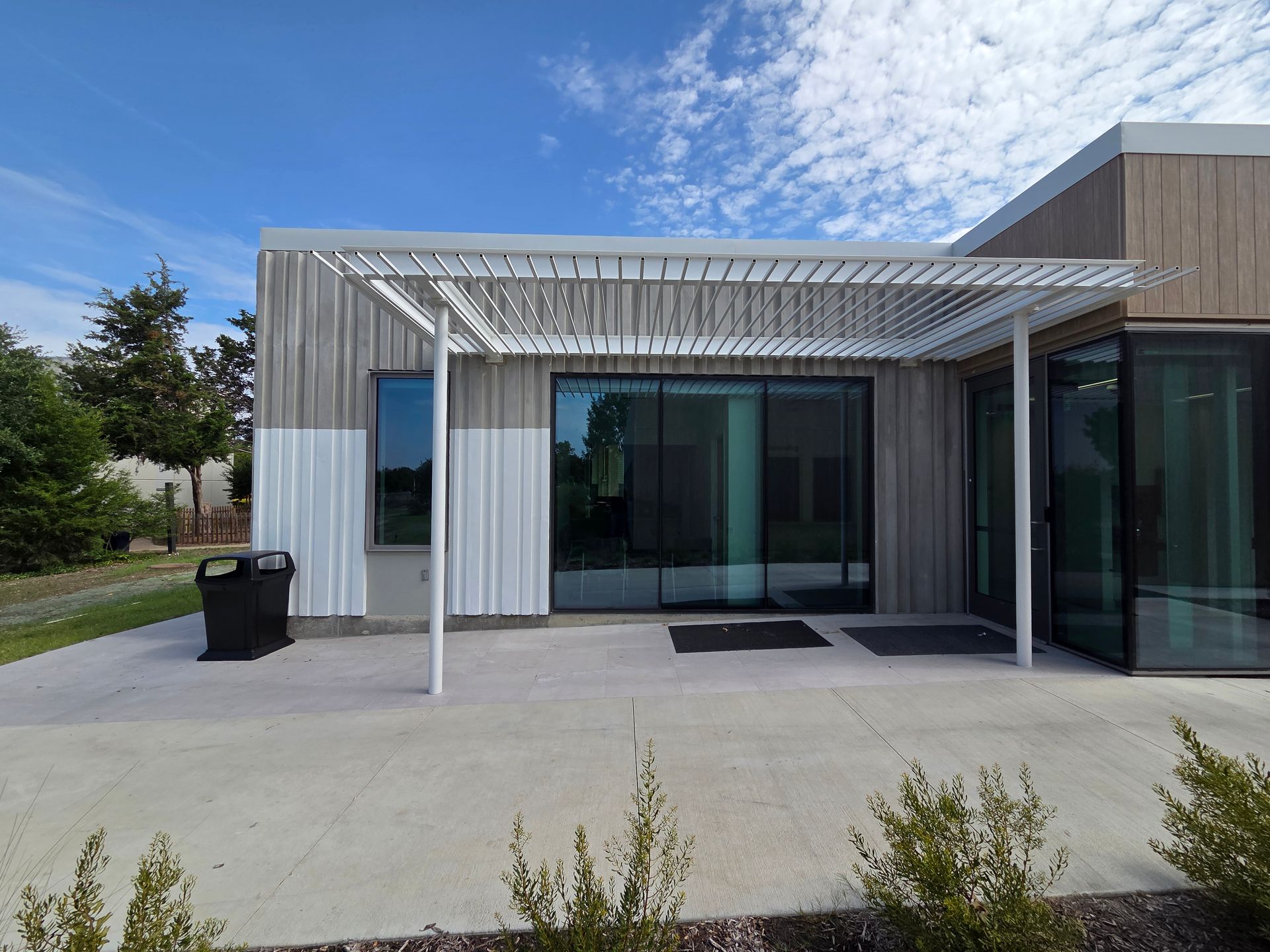 Entrance to a modern building with a white pergola, glass doors, and a concrete patio on a sunny day.