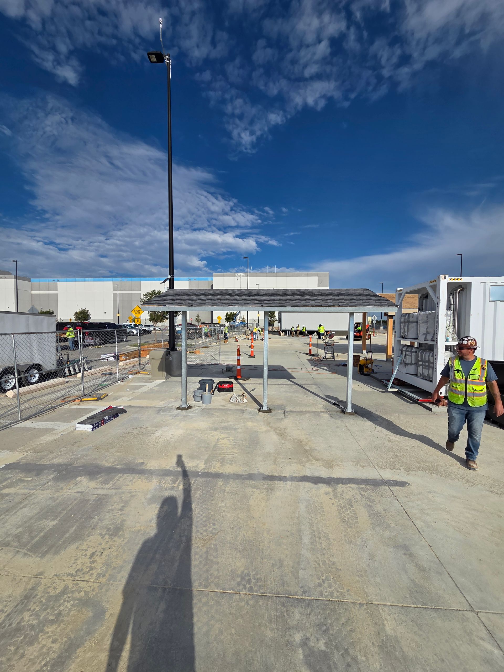 Construction site: solar panel canopy, worker walking, tall light pole, bright blue sky.