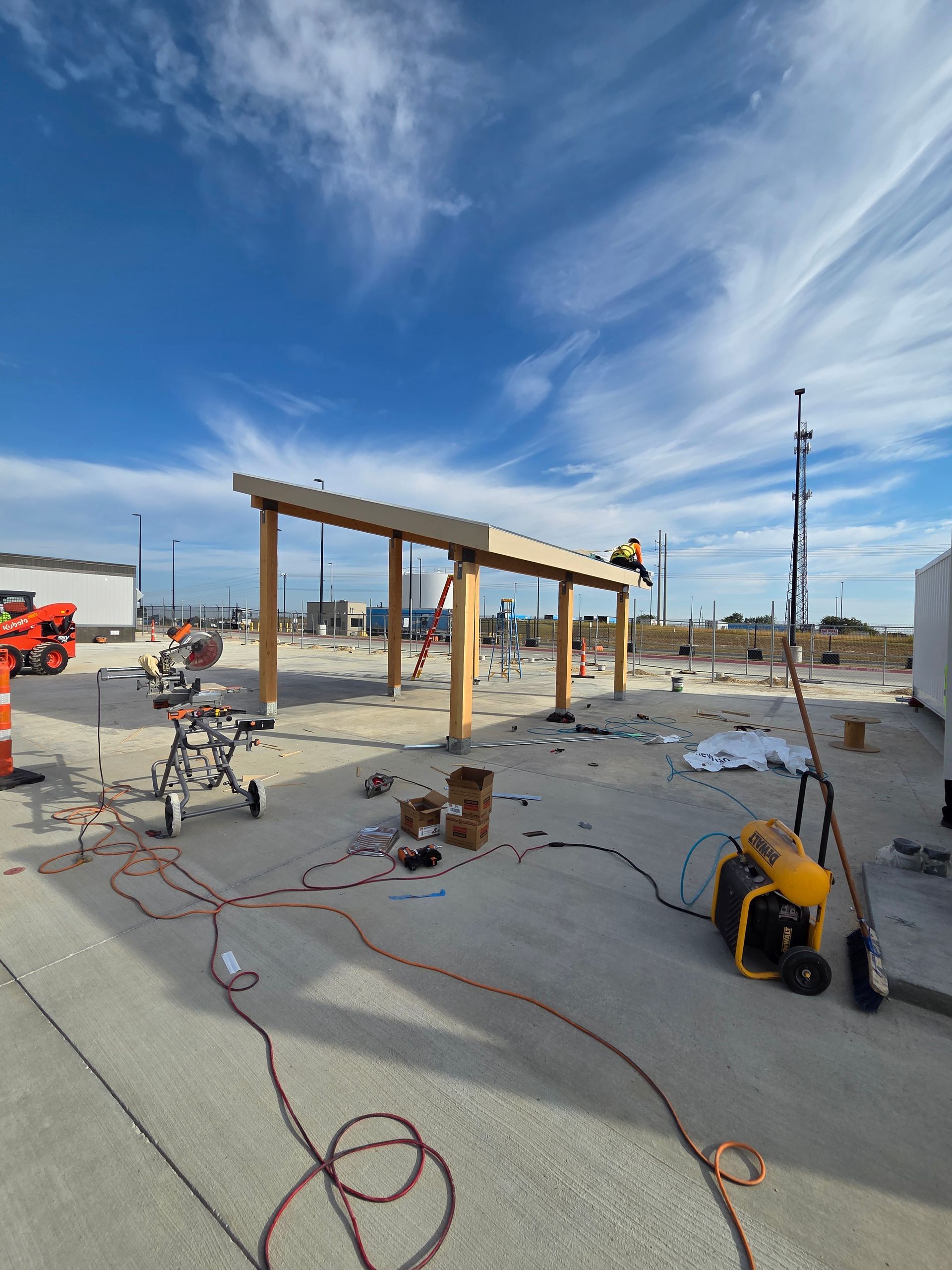Construction site with wooden structure being built under a cloudy blue sky. Workers and tools are visible.