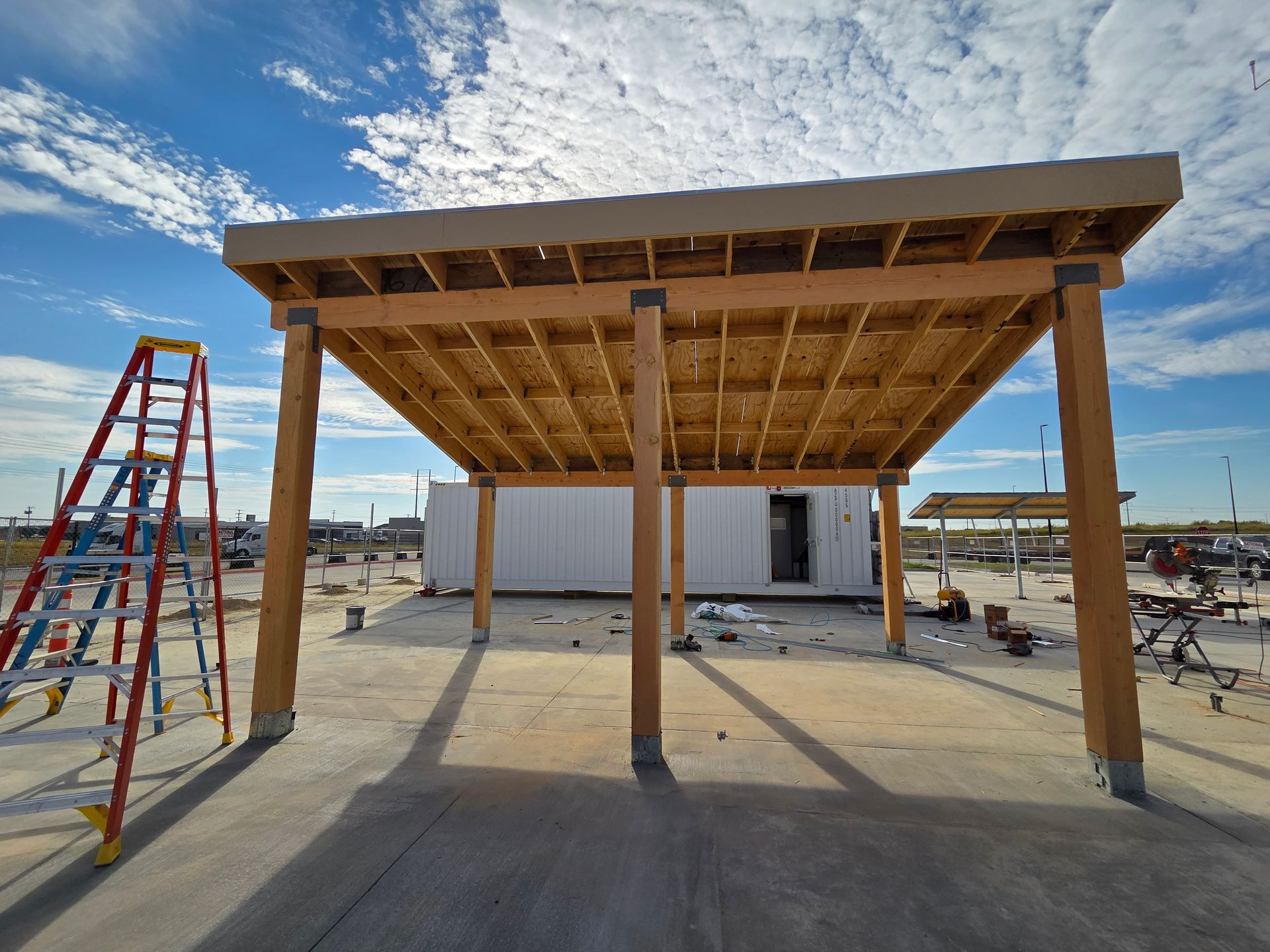 Wooden carport under construction on a concrete slab, with a ladder and blue sky in background.