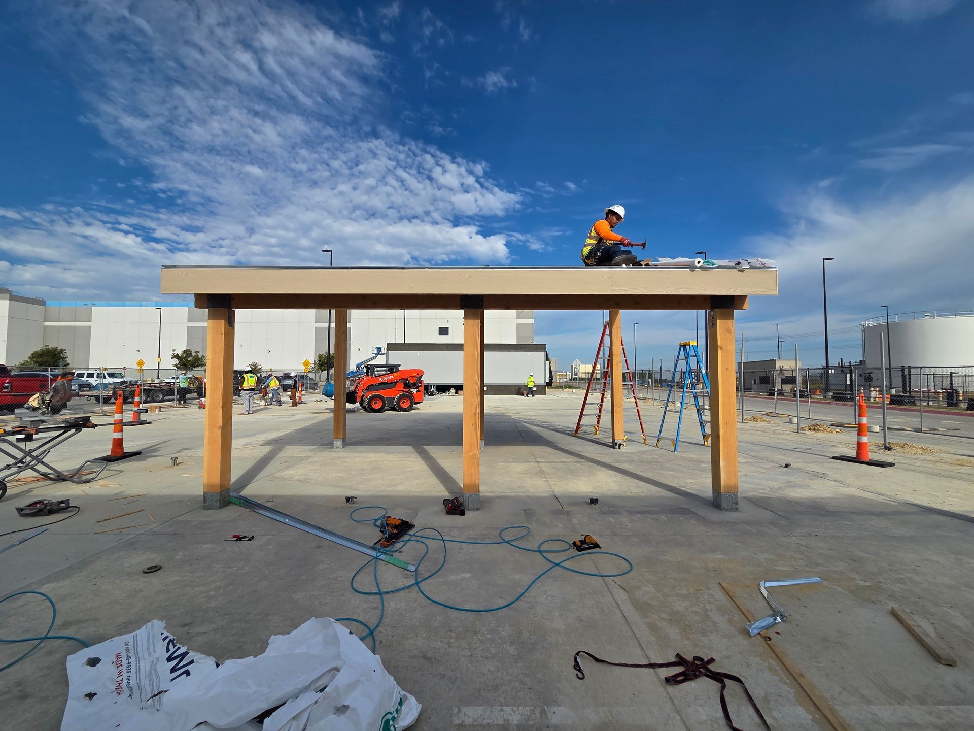 Construction worker on a wooden shelter roof, other workers nearby. Industrial setting, blue sky.