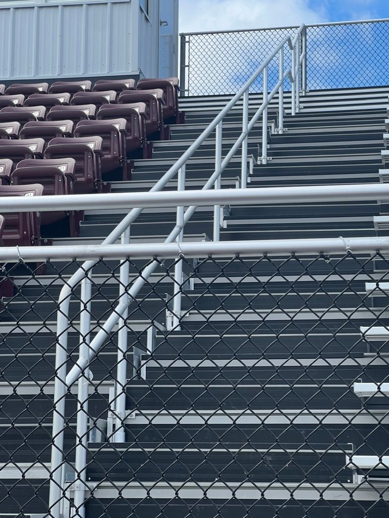 Bleachers with maroon seats, stairs with railing, chain-link fence, and a cloudy sky.