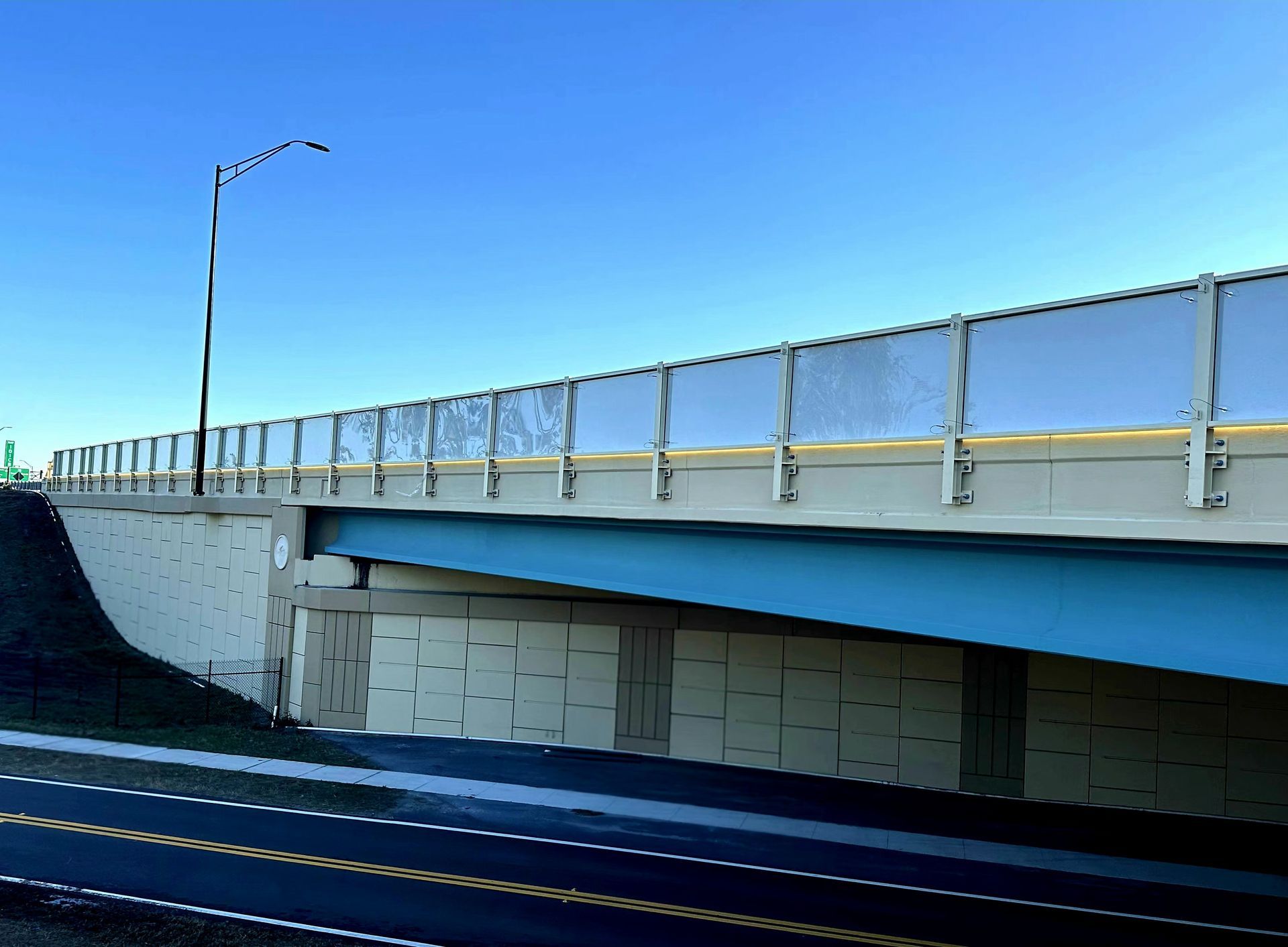 Bridge with clear barrier and blue underside against a blue sky.
