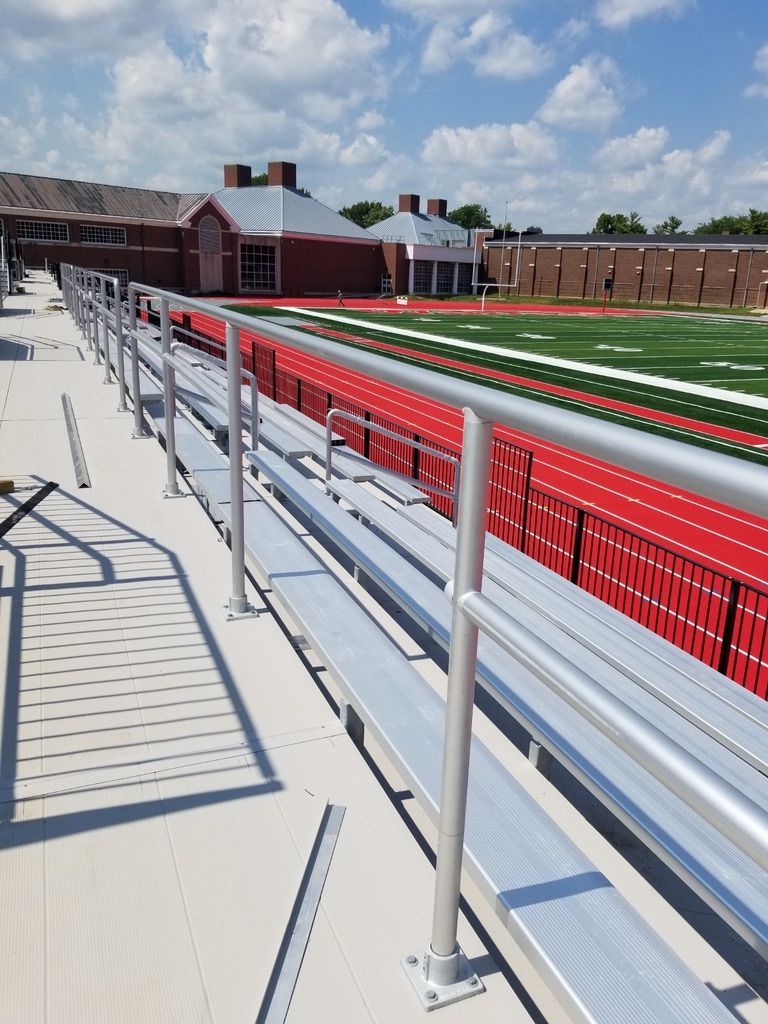 Bleachers overlooking a red track and green field. A building with red brick and a white roof is in the background.